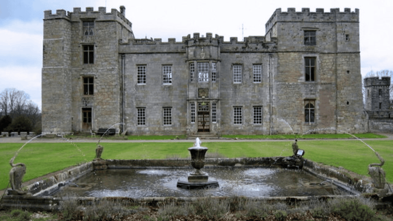 A wide, straight-on shot of Chillingham Castle, a historic stone fortress with a square water fountain spraying water in the green foreground lawn.